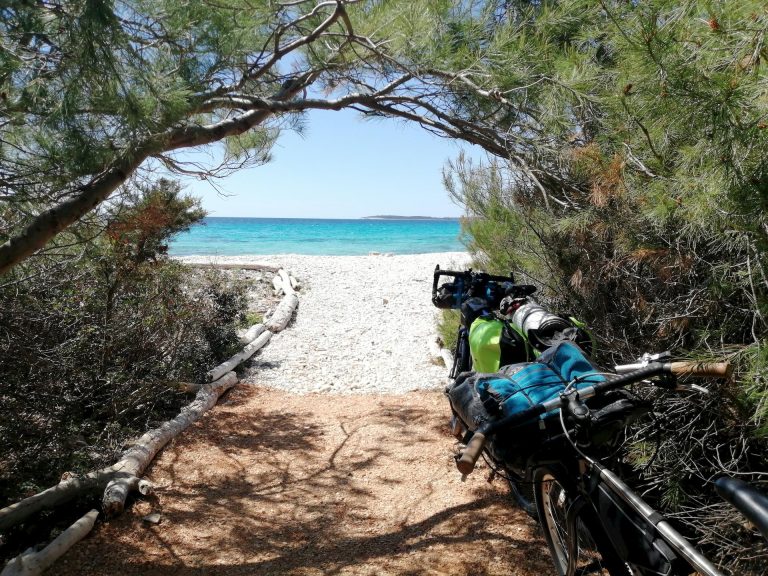 White pebble path through the pine woods leading to the beach with a bike parked on the right side.