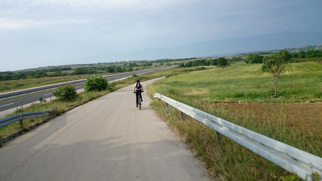 A girl on a bike, from behind, cycling on an empty road surrounded by green meadows. Zadar cycling terrain.